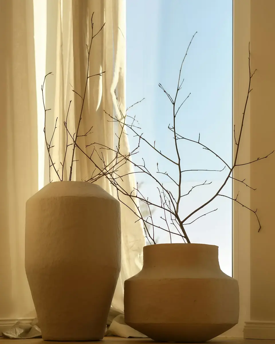 Vasos de cerâmica com galhos secos junto a uma janela com cortina clara